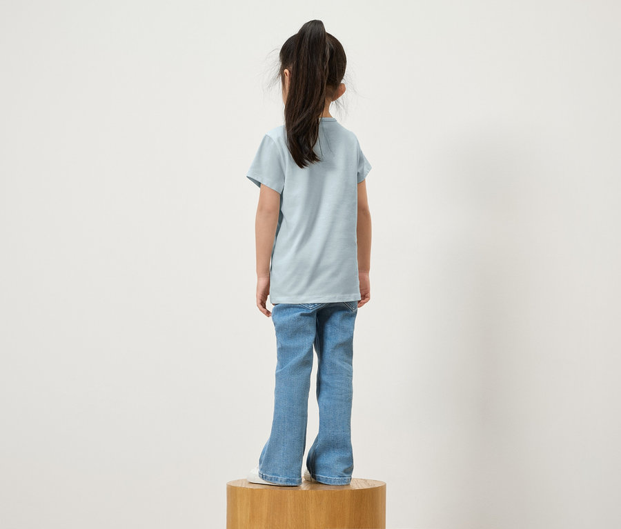 Une fille avec une queue de cheval se tient de dos sur un socle en bois, portant un t-shirt bleu et un jean.