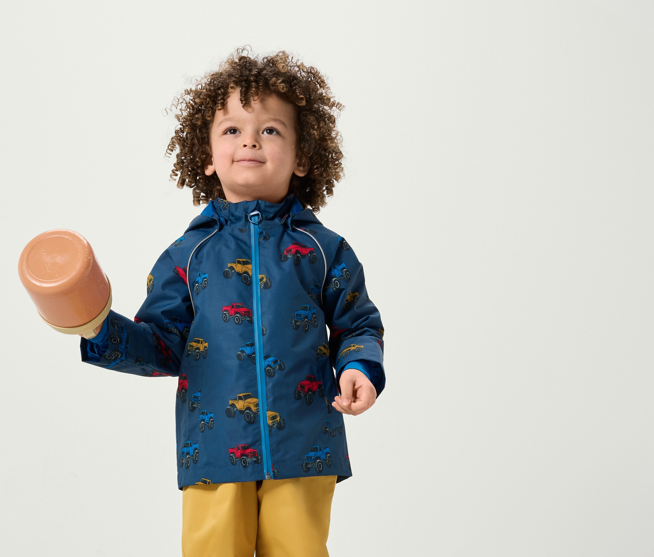 Un garçon aux cheveux bouclés tient un récipient marron. Il porte une veste avec un imprimé de voiture et un pantalon jaune.
