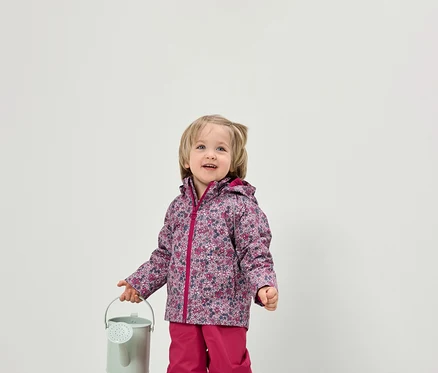 Un jeune enfant tient un petit arrosoir vert. Il porte une veste à fleurs et un pantalon rose.