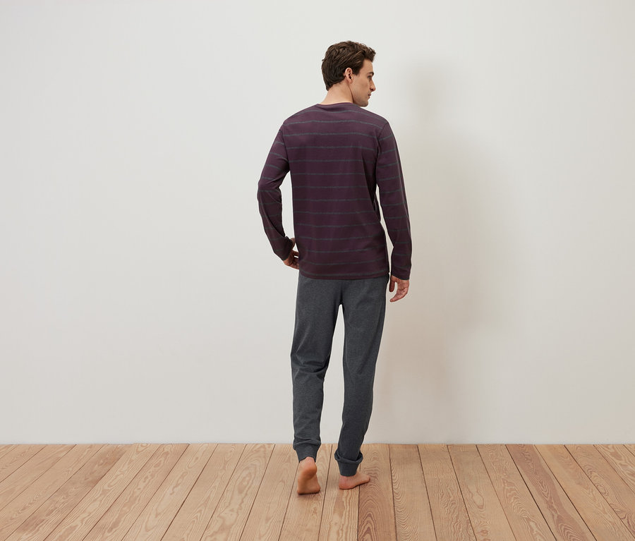 Un homme vu de dos, vêtu d'un t-shirt à manches longues rayé et d'un pantalon de jogging gris, se tient debout sur un plancher en bois.