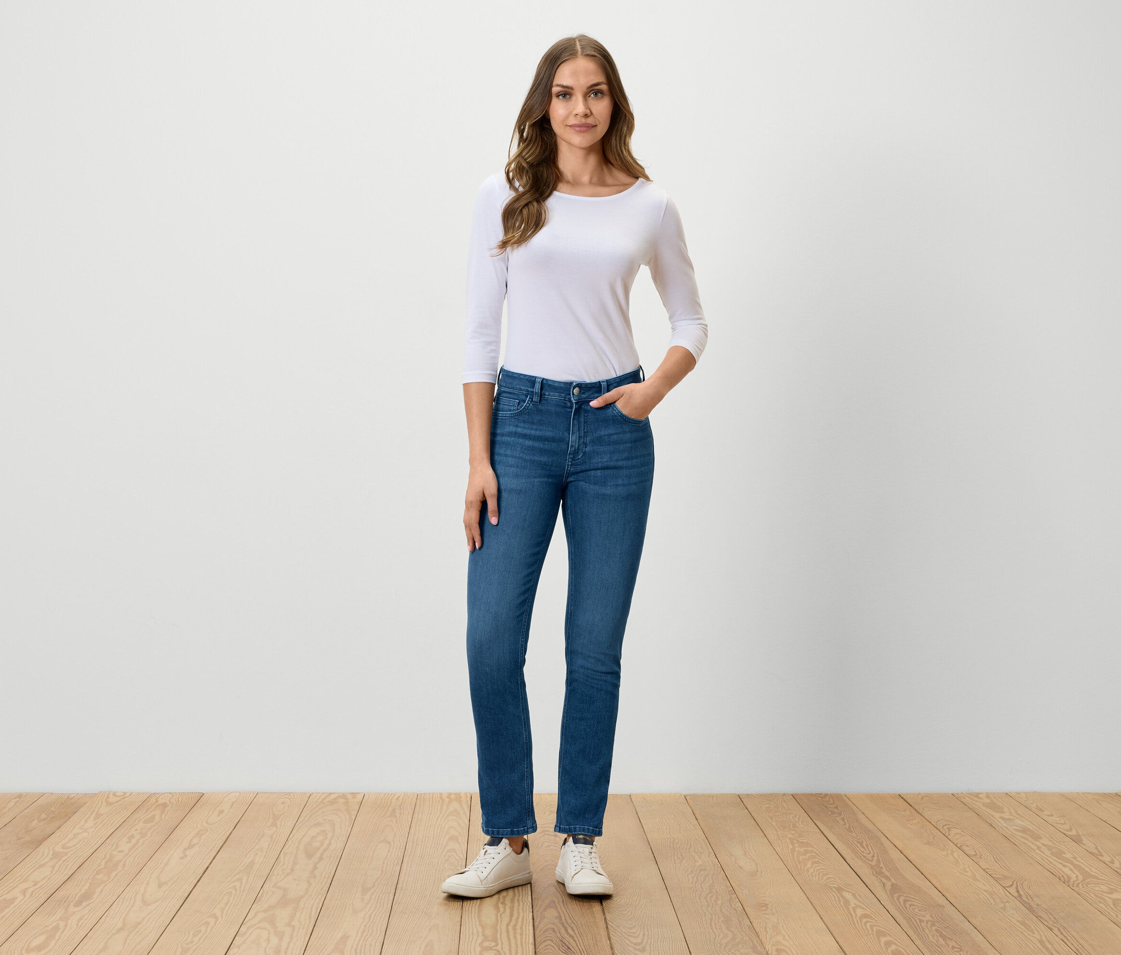 Une femme se tient debout et pose en jean bleu, t-shirt blanc et baskets blanches.