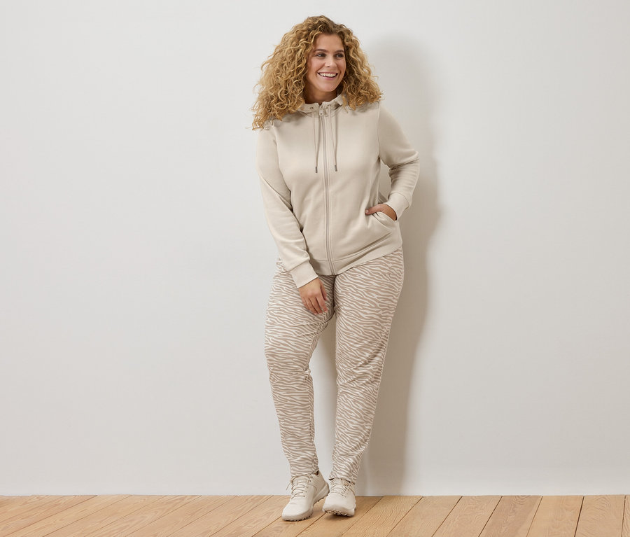 Femme aux cheveux bouclés portant un sweat à capuche beige et un pantalon à imprimé zébré, appuyée contre un mur blanc.