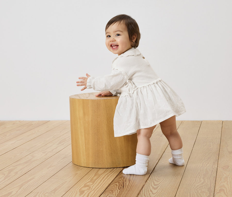 Un bébé souriant en robe blanche se tient à côté d'un petit tabouret en bois sur un plancher en bois.
