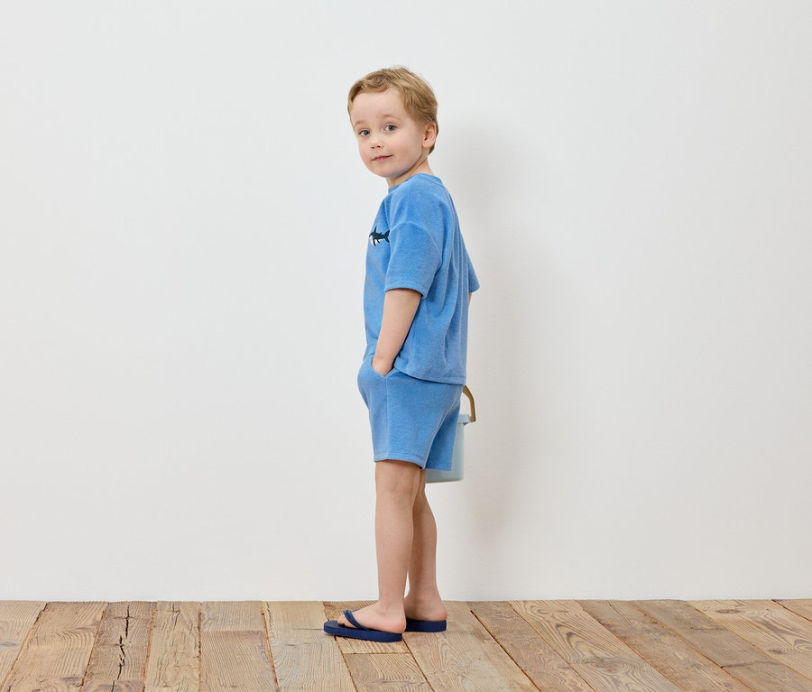 Un garçon blond se tient les mains dans les poches devant un mur blanc, vêtu d'un t-shirt et d'un short bleu clair, avec des tongs bleues.