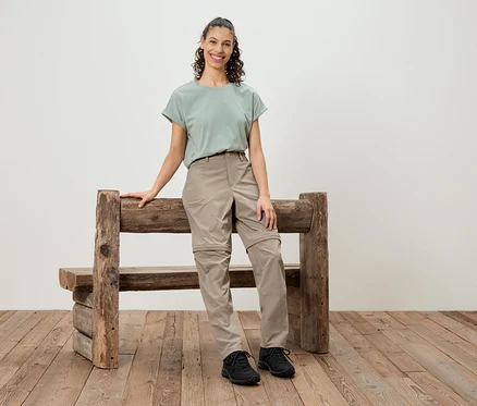 Une femme se tient à côté d'un banc en bois rustique, portant un t-shirt vert, un pantalon beige et des chaussures noires.