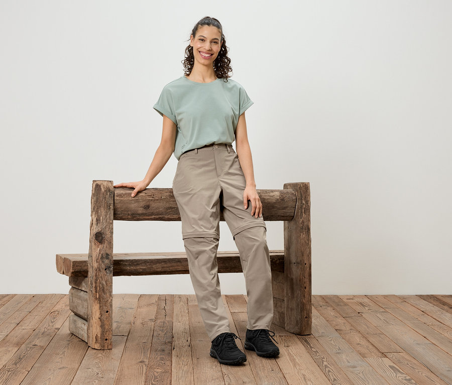 Une femme se tient à côté d'un banc en bois rustique, portant un t-shirt vert, un pantalon beige et des chaussures noires.