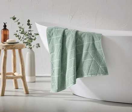 Une serviette vert menthe à motif géométrique est suspendue au bord d'une baignoire blanche. Un tabouret en bois avec du savon et un distributeur est à côté.