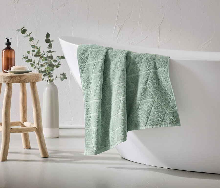 Une serviette vert menthe à motif géométrique est suspendue au bord d'une baignoire blanche. Un tabouret en bois avec du savon et un distributeur est à côté.
