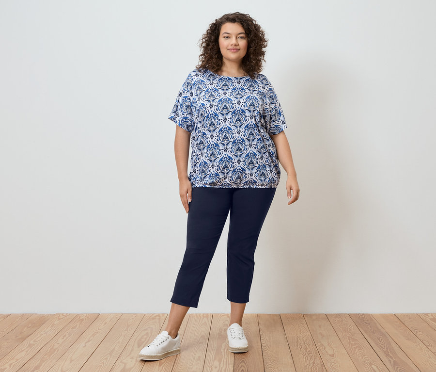 Une femme aux cheveux bouclés se tient devant un mur, portant un chemisier à motifs bleus, un pantalon bleu marine et des baskets blanches.