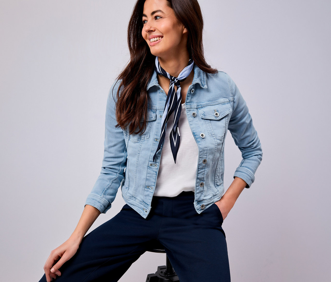 Une femme souriante avec un foulard bleu et blanc rayé, une veste en jean et un pantalon bleu est assise sur un tabouret.
