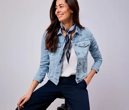 Une femme souriante avec un foulard bleu et blanc rayé, une veste en jean et un pantalon bleu est assise sur un tabouret.