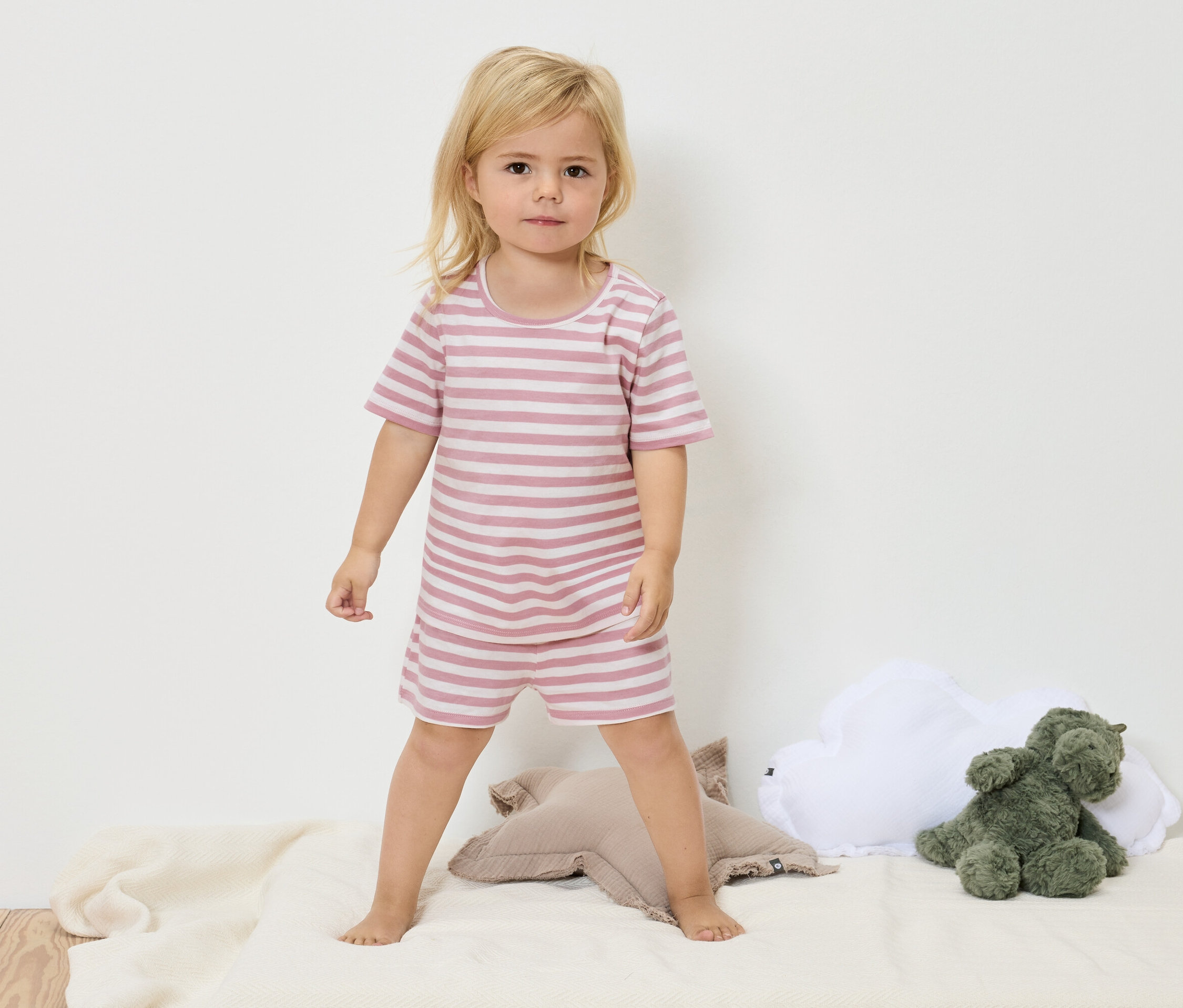 Une petite fille blonde se tient debout sur une couverture couleur crème et porte un pyjama rayé rose et blanc.