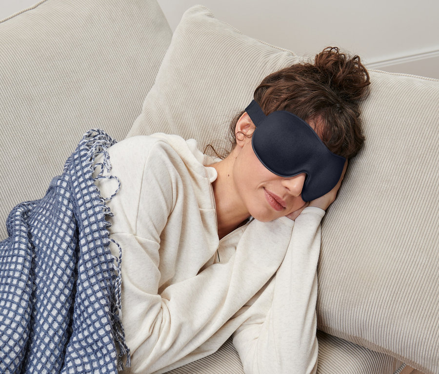 Une femme aux cheveux bruns relevés portant un masque de sommeil est allongée sur un canapé beige sous une couverture bleu et blanc.