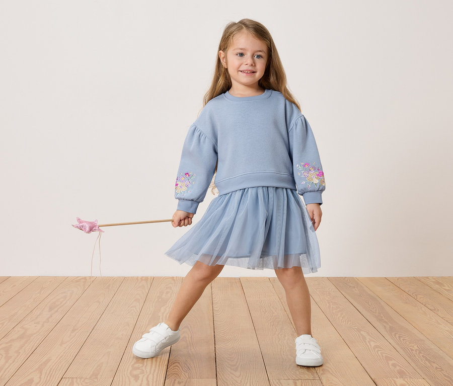 Fillette portant un pull bleu avec des broderies florales et une jupe en tulle, tenant une baguette magique.