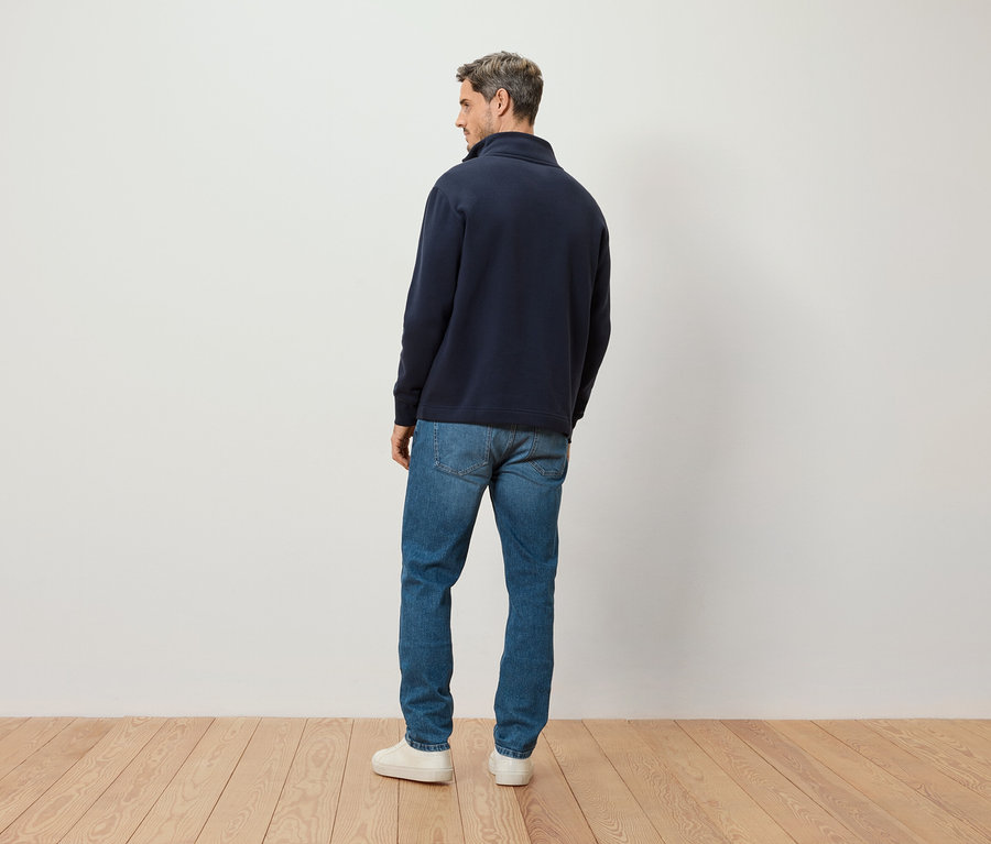 Un homme se tient dos à la caméra, portant un pull bleu marine zippé, un jean et des baskets blanches.