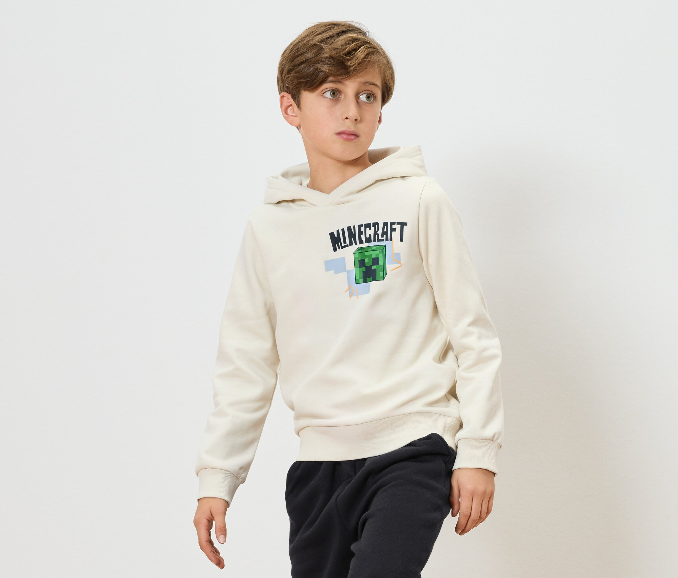 Un garçon porte un sweat à capuche Minecraft crème et un jogging noir sur fond blanc.