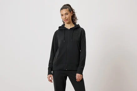 Une femme porte un survêtement noir composé d'un sweat à capuche zippé et d'un pantalon sur un fond neutre.