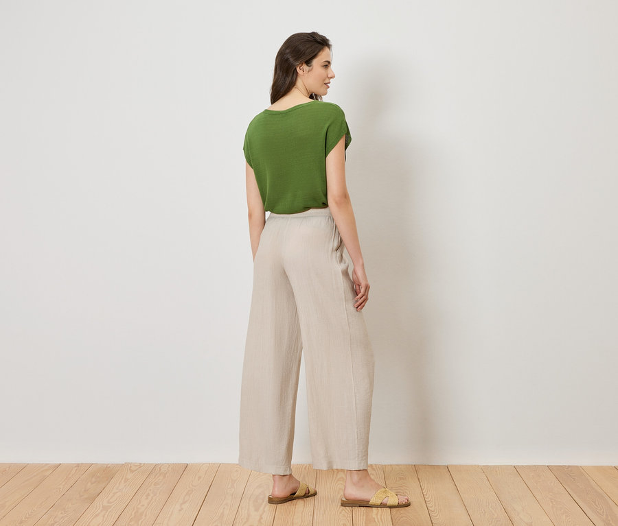 Une femme se tient dos à la caméra, vêtue d'un t-shirt vert, d'un pantalon beige et de sandales jaunes.