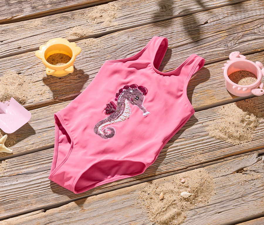 Un maillot de bain rose avec un motif d'hippocampe est posé sur un plancher en bois, entouré de sable, d'une pelle, d'une étoile de mer et de deux moules à sable.