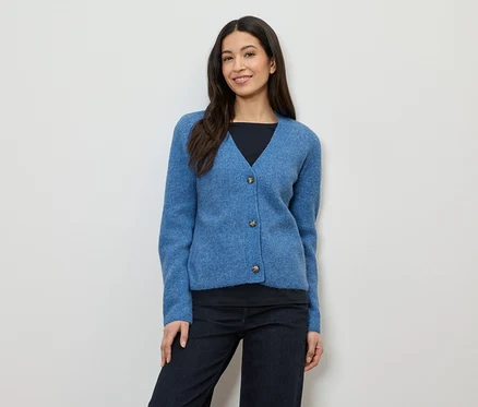 Une femme aux cheveux foncés porte un cardigan bleu à trois boutons sur un haut noir et un pantalon foncé.