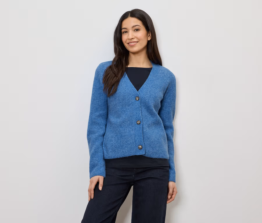 Une femme aux cheveux foncés porte un cardigan bleu à trois boutons sur un haut noir et un pantalon foncé.