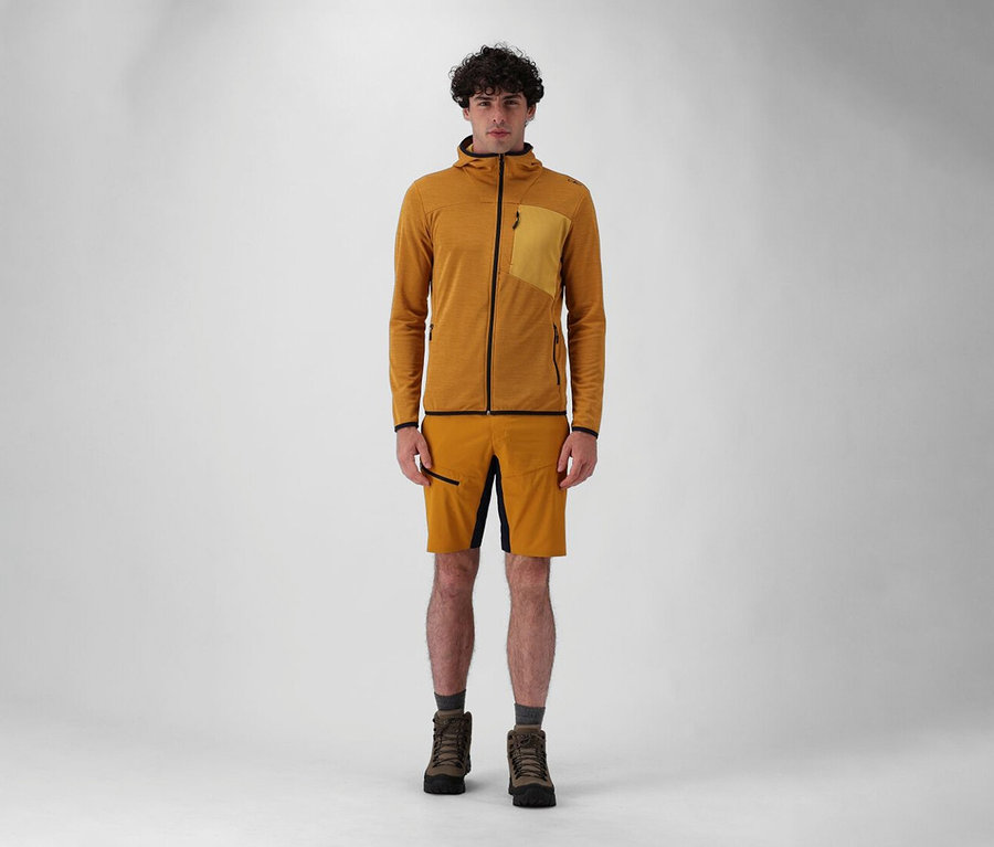 Un homme debout en short et bottes portant une veste à capuche CMP pour homme couleur moutarde sur fond blanc.