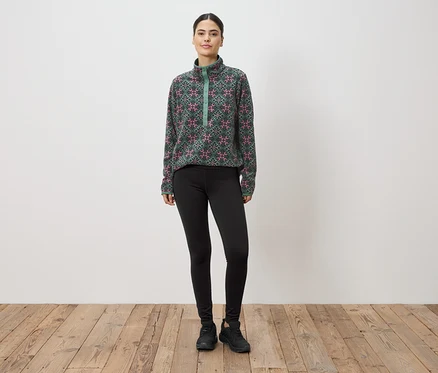 Une femme se tient devant un mur, portant un haut en polaire à motifs verts et roses, des leggings et des baskets.
