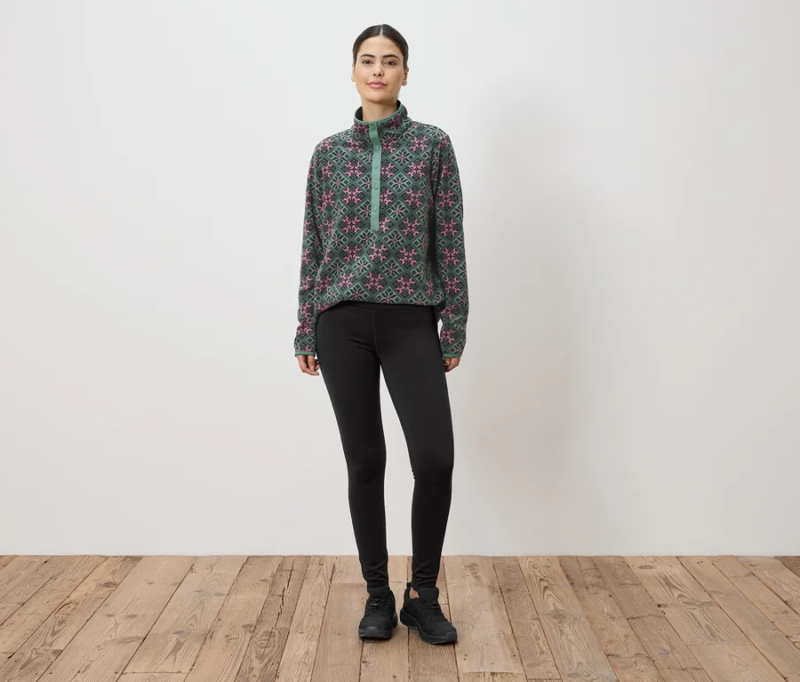 Une femme se tient devant un mur, portant un haut en polaire à motifs verts et roses, des leggings et des baskets.