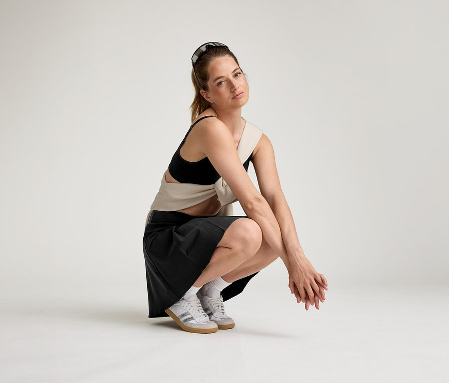 Une femme portant des lunettes de soleil sur la tête est accroupie sur un fond blanc, vêtue d'un soutien-gorge de sport noir, d'un haut beige, d'une jupe noire et de baskets blanches.