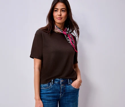 Une femme portant un T-shirt Street One marron foncé et un jean bleu, avec un foulard rose à imprimé animal autour du cou, se tient la main dans la poche.