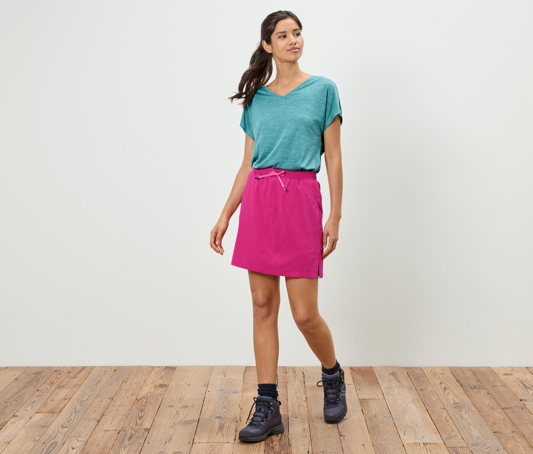 Une femme marche sur un plancher en bois devant un mur blanc, vêtue d'un t-shirt turquoise, d'une jupe rose et de chaussures de randonnée.