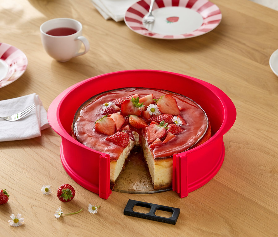Cheesecake aux fraises avec cercle à gâteau rouge, coupé, sur table en bois avec marguerites, couverts et boissons.