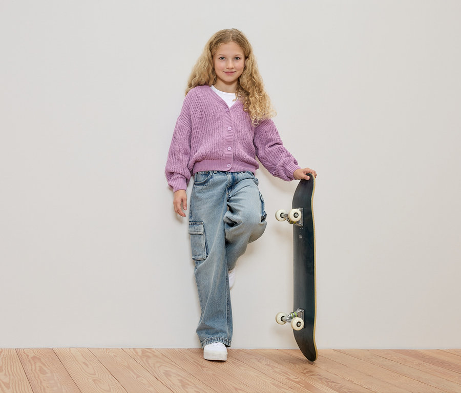 Une fille aux cheveux blonds bouclés, portant un cardigan violet et un jean, appuyée contre un skateboard devant un mur blanc.