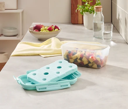 Sur un comptoir de cuisine, on trouve un bol de fruits, un linge jaune, un récipient de fruits coupés et un bloc réfrigérant dans une boîte.