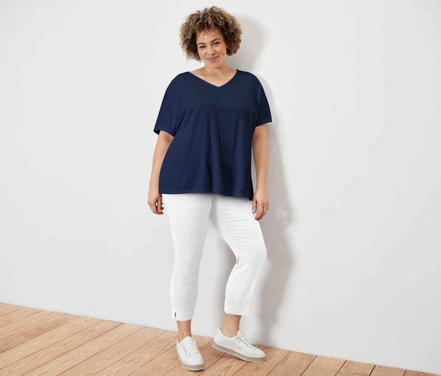 Femme aux cheveux bouclés portant un t-shirt bleu marine à col en V, un pantalon blanc et des chaussures crème, appuyée contre un mur blanc.