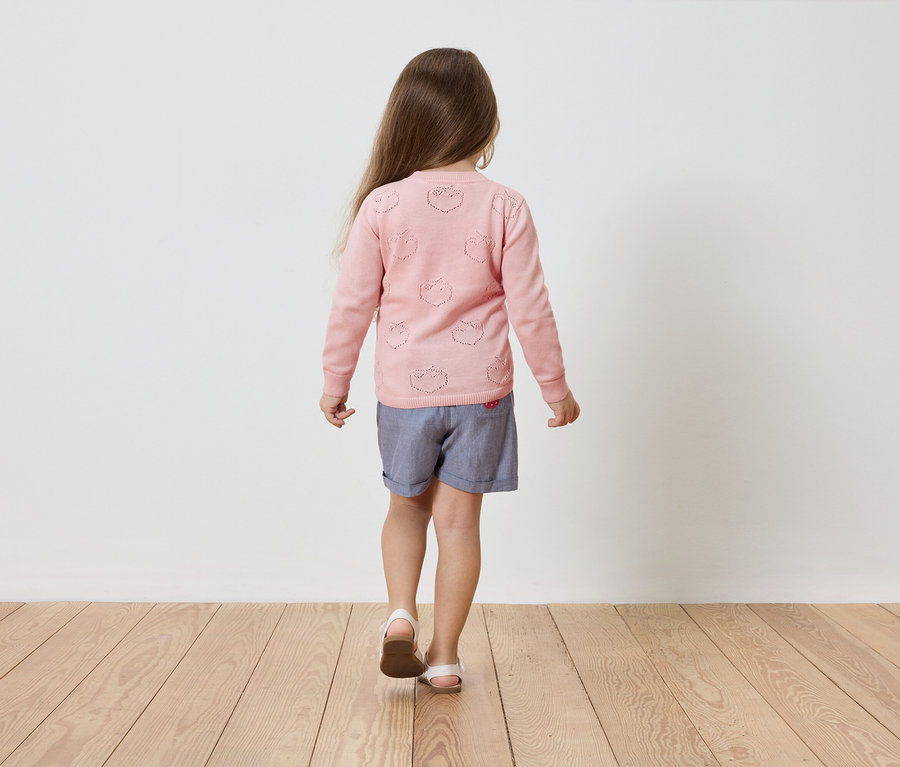 Fillette vue de dos portant un cardigan rose à motifs ajourés de fraises, un short bleu et des sandales sur un plancher en bois.