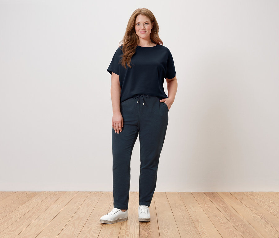 Une femme se tient devant un mur, portant un t-shirt bleu marine, un pantalon bleu marine et des chaussures blanches.