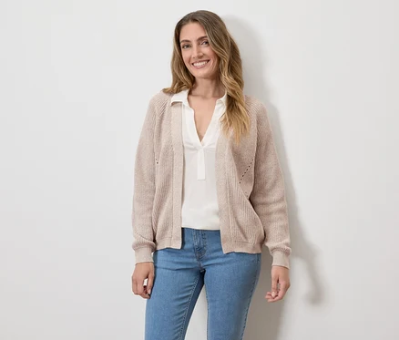Une femme souriante portant un cardigan beige, un chemisier blanc et un jean devant un mur blanc.