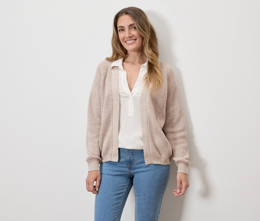 Une femme souriante portant un cardigan beige, un chemisier blanc et un jean devant un mur blanc.