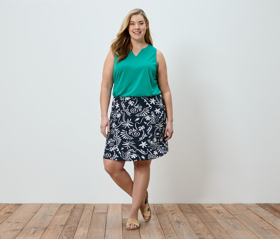 Une femme se tient devant un mur blanc, portant un haut vert et une jupe bleu marine à motifs blancs.