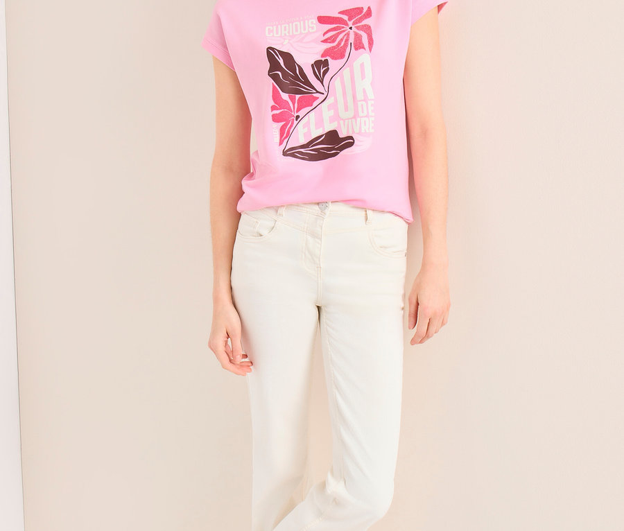 Une femme souriante, portant une casquette beige et un jean clair, est appuyée contre un mur. Elle porte un T-shirt à motif CECIL rose avec un imprimé floral.