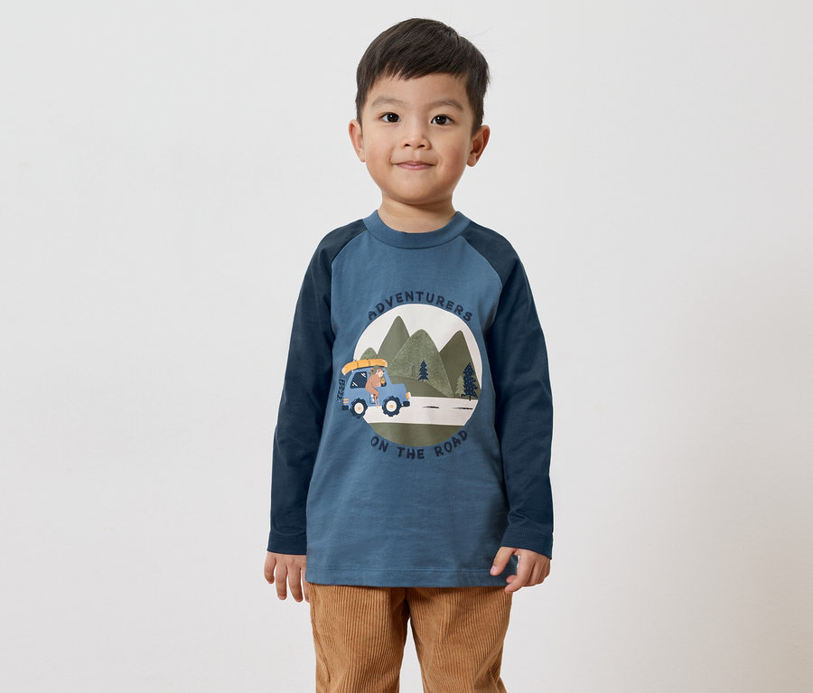 Un garçon portant un t-shirt bleu à manches longues avec un motif d'ours en voiture et un pantalon en velours côtelé marron sur fond blanc.