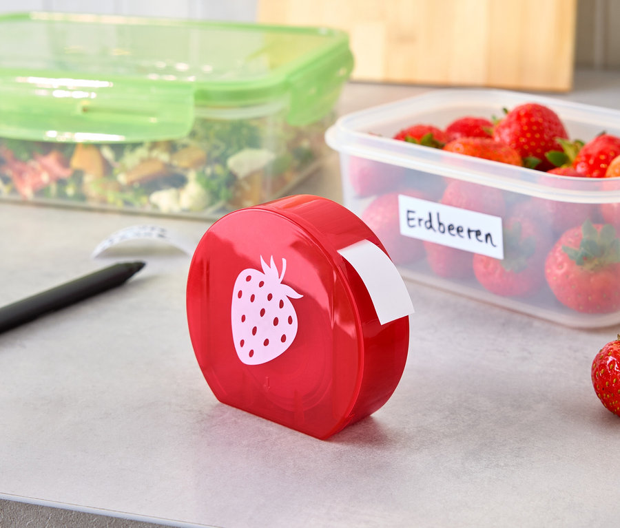 Un distributeur d'étiquettes rouge avec un motif de fraise se trouve à côté d'une boîte de fraises étiquetée et d'une boîte de salade sur une surface grise.