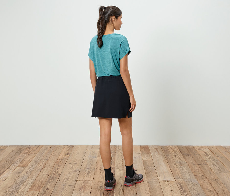 Une femme se tient dos à la caméra, portant un t-shirt bleu-vert, une jupe noire et des chaussures de sport.