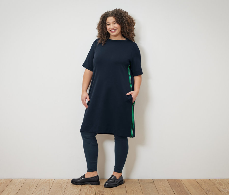 Une femme aux cheveux bouclés porte une robe bleu marine à rayures vertes, un legging bleu marine et des mocassins noirs.
