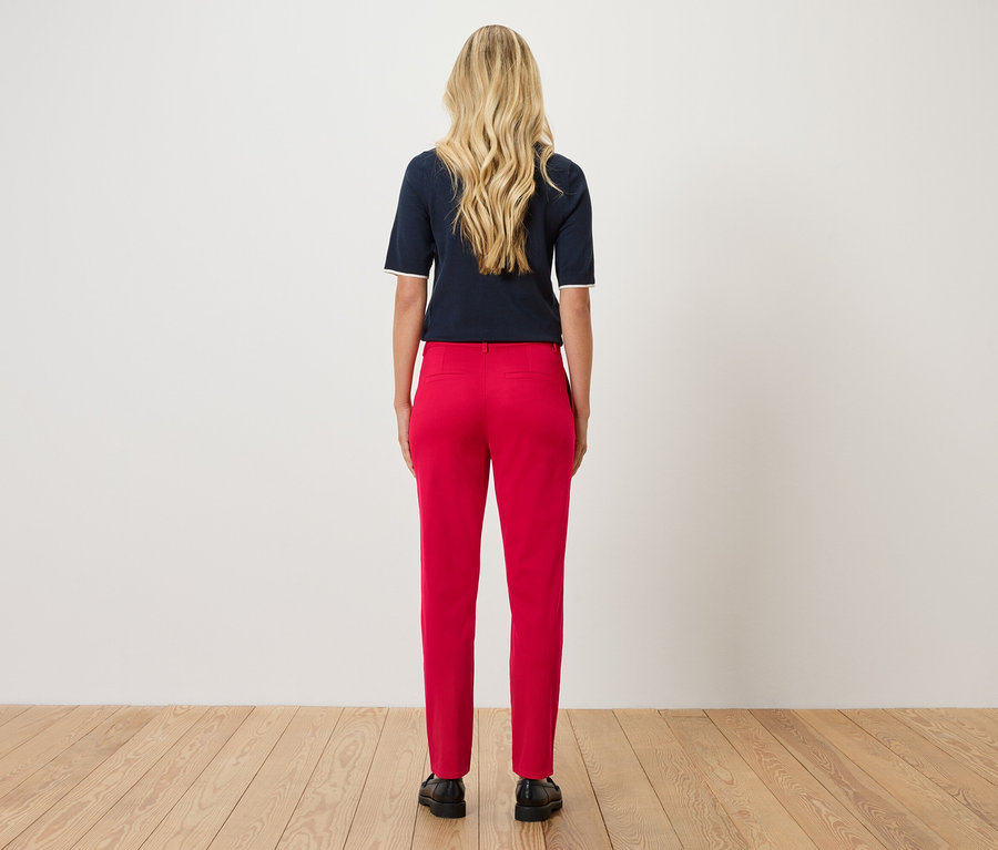 Une femme aux cheveux blonds et ondulés se tient dos à la caméra, portant un haut bleu marine et un pantalon rouge.