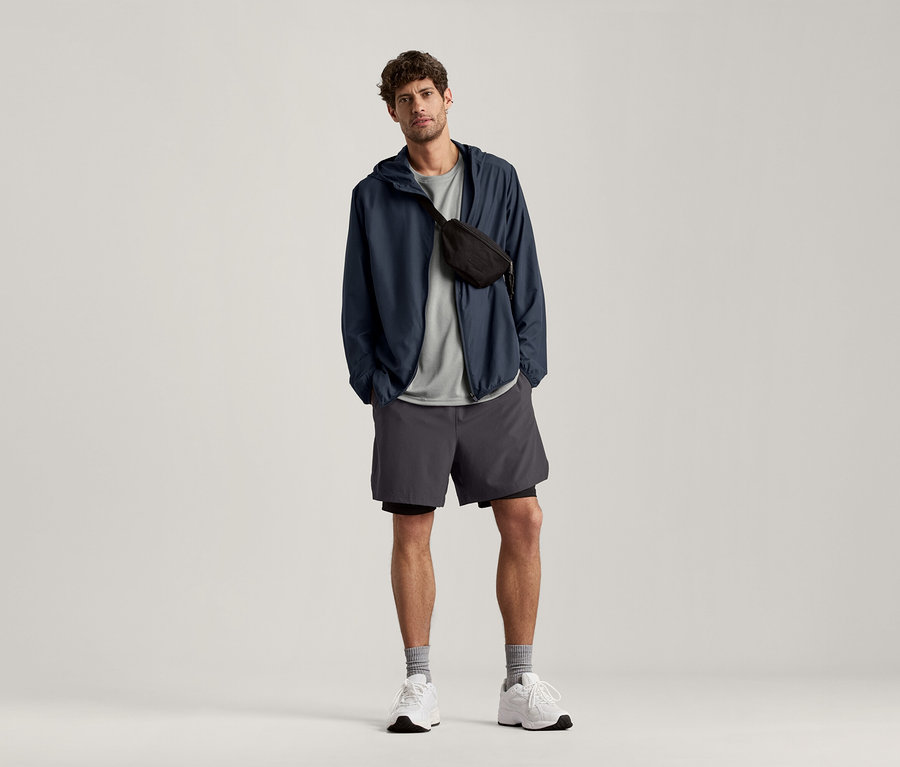 Un homme debout porte un t-shirt de sport gris clair chiné, une veste bleu marine et un short gris.