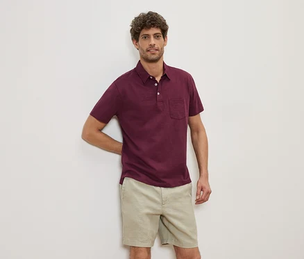 Un homme est appuyé contre un mur et porte un polo bordeaux et un short beige.