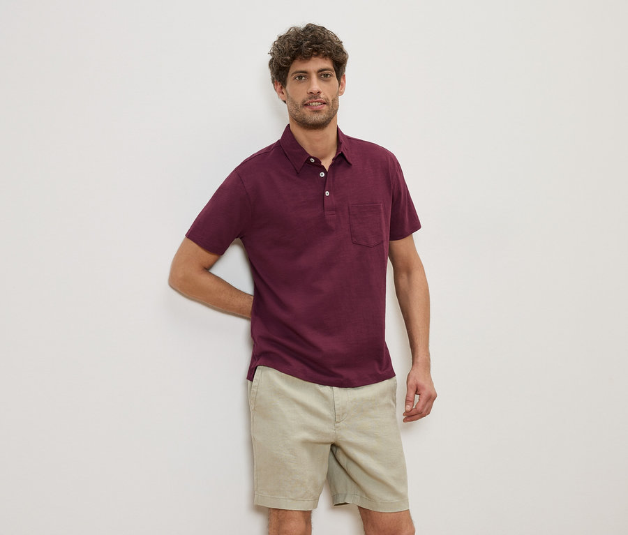 Un homme est appuyé contre un mur et porte un polo bordeaux et un short beige.