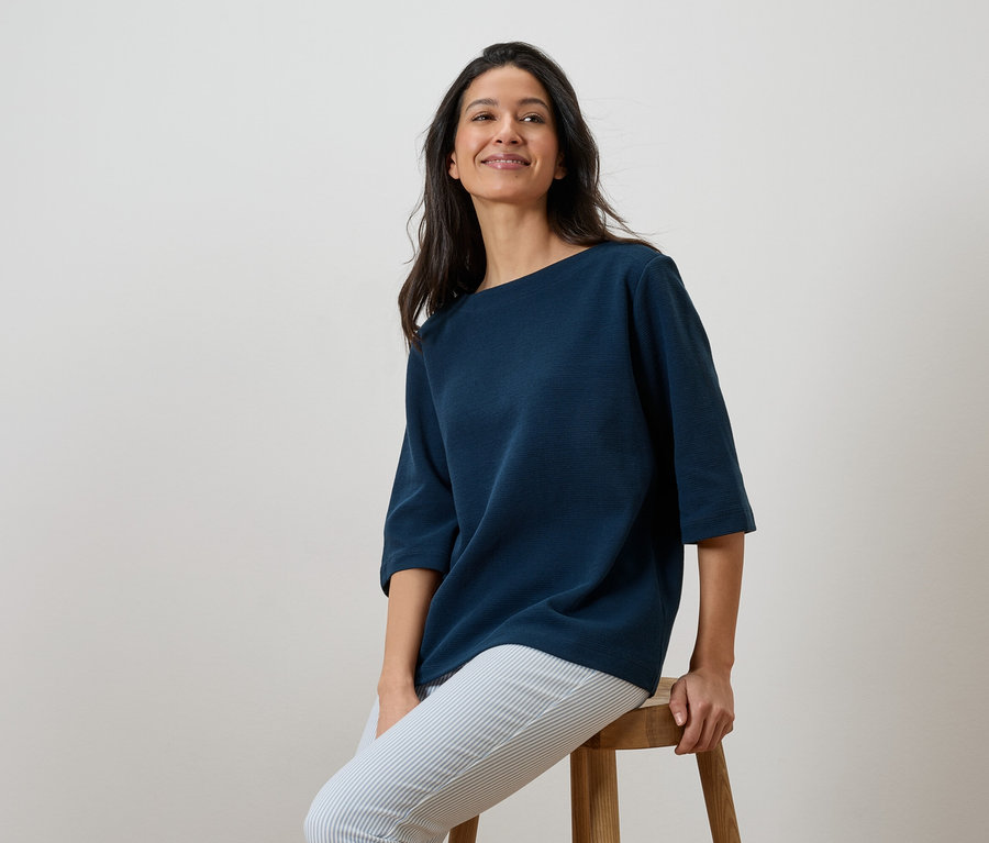 Femme assise sur un tabouret, portant un t-shirt bleu marine et un pantalon rayé.